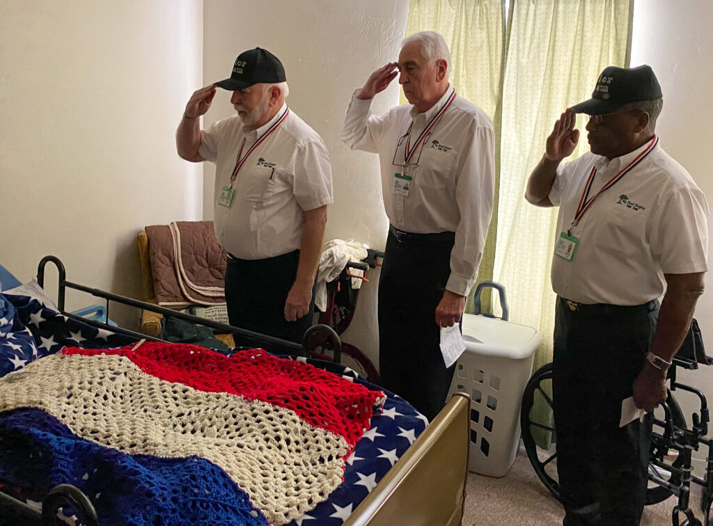 Three Valor Team members saluting an unseen Veteran Patient in bed covered with a red white and blue afgan.